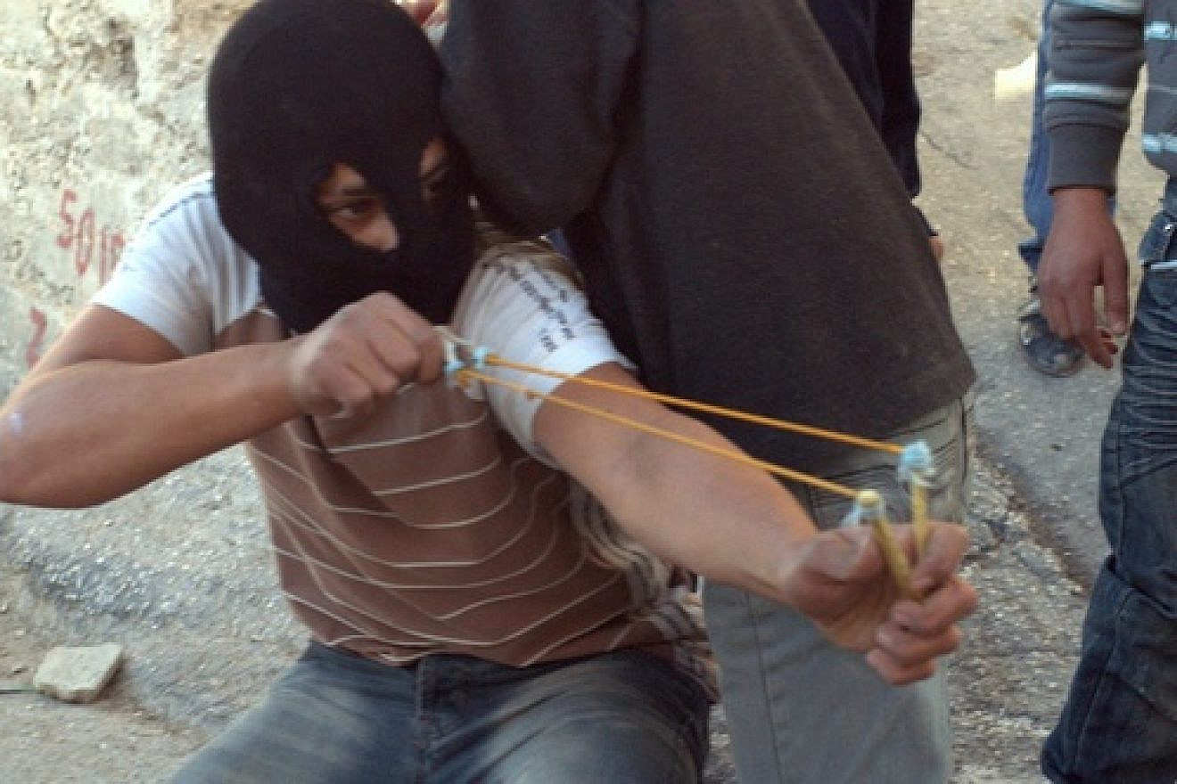 Click photo to download. Caption: In Silwan, an older balaclava-clad child with a slingshot fires off a stone at the cops, as cameras in front of him captured his “heroic” pose. Credit: B. Davidson.