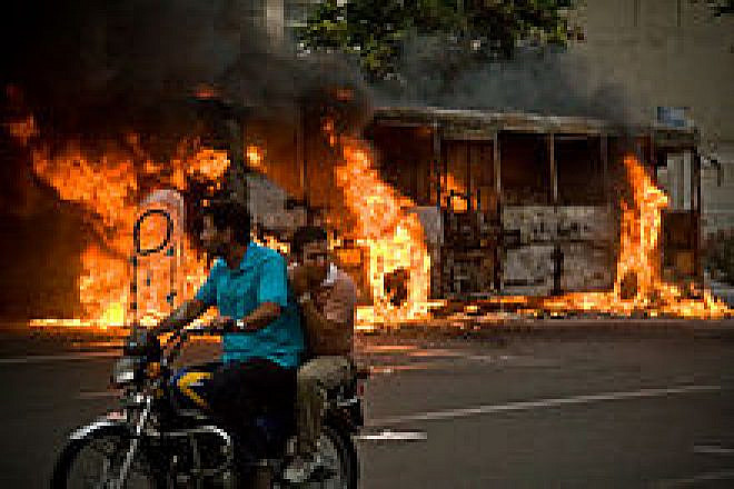 A bus burns on the streets of downtown Tehran as protesters cycle by in June 2018. Credit: Wikipedia.