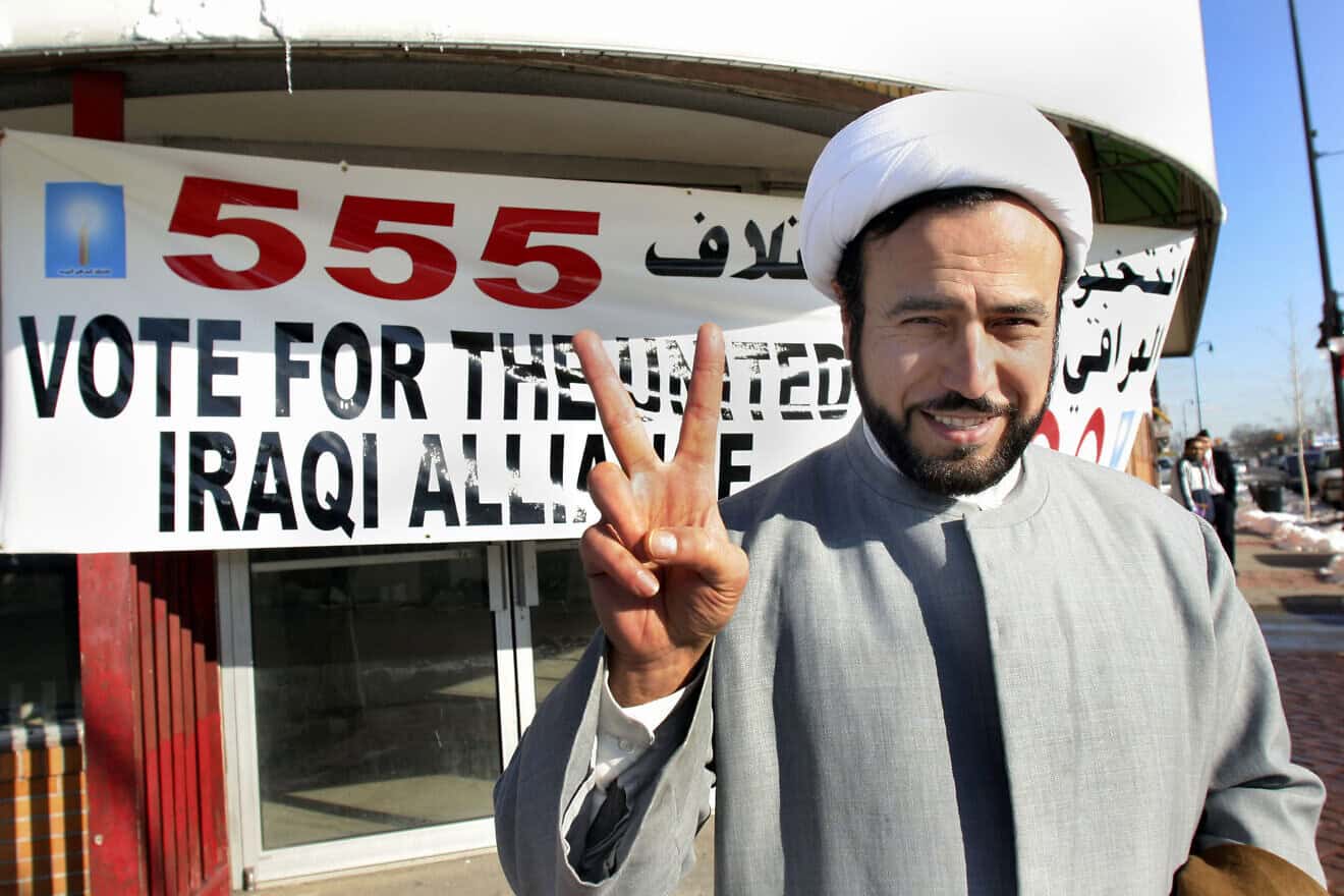 Imam Husham al-Husainy of the Karbaala Islamic Institute of America makes the peace sign with his fingers while standing in front of a "Vote for the Iraqi United Alliance" banner on Dec. 12, 2005 in Dearborn, Michigan. Photo by Bill Pugliano/Getty Images.